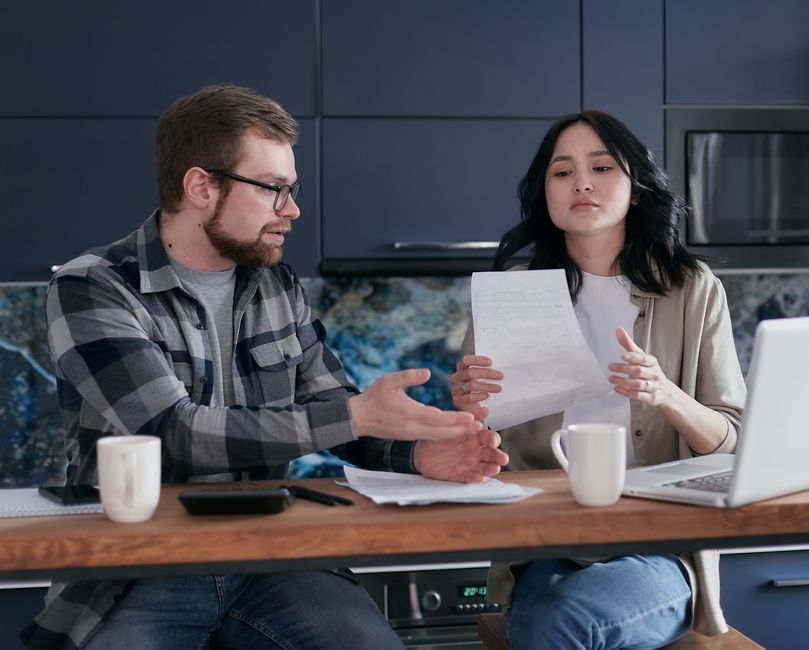 A couple stands in a modern kitchen, leaning against the counter with serious expressions, conveying the financial and emotional weight of a major life decision.